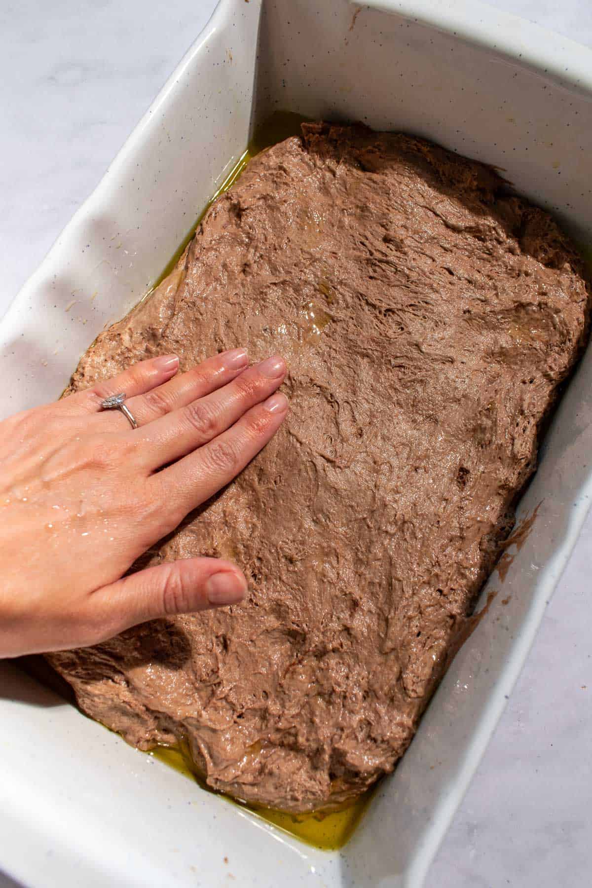A hand pressing chocolate bread dough into a large white baking dish.