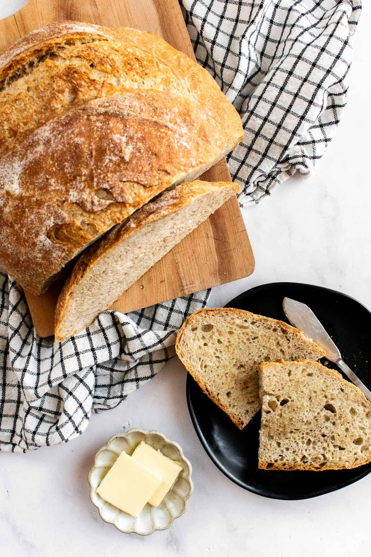 Two small slices of crusty bread on a black plate next to a dish of butter squares and the rest of the loaf on a wooden board.