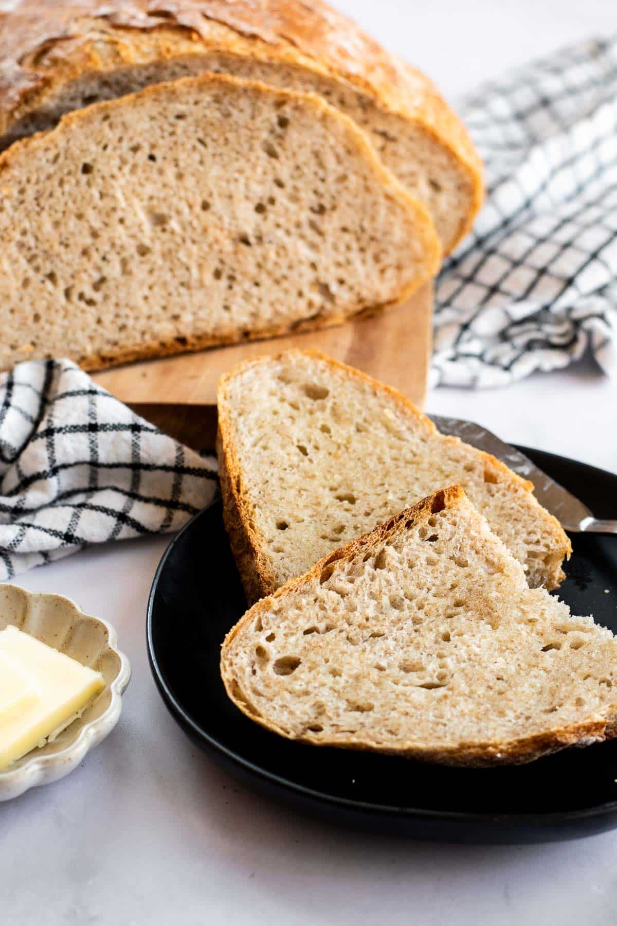 Two slices of crusty bread on a black plate next to a dish of butter squares and a rustic loaf on a wood board.