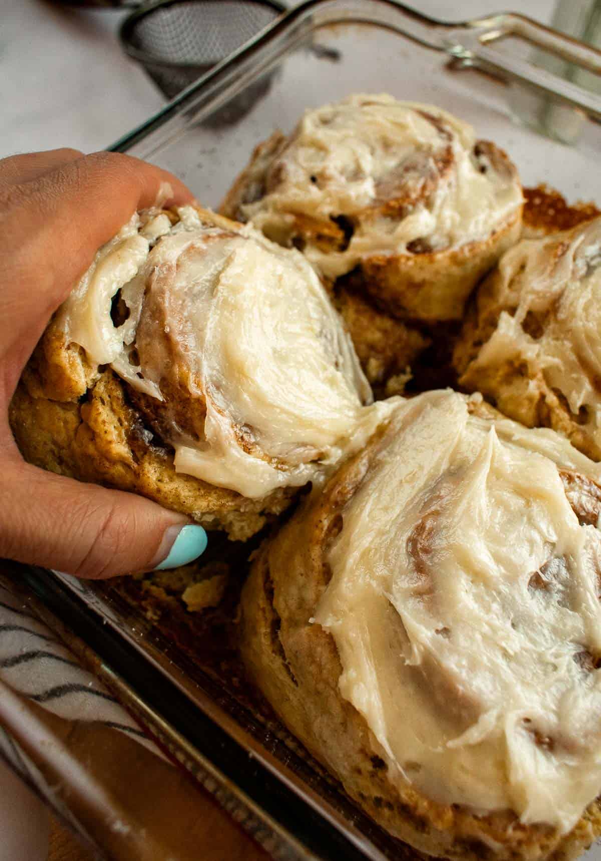 A hand lifting a frosted cinnamon roll out of a glass baking dish with three more cinnamon rolls.
