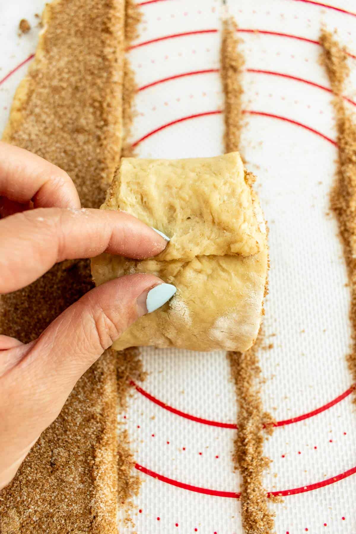 A hand pinching the end of a rolled-up unbaked cinnamon roll.