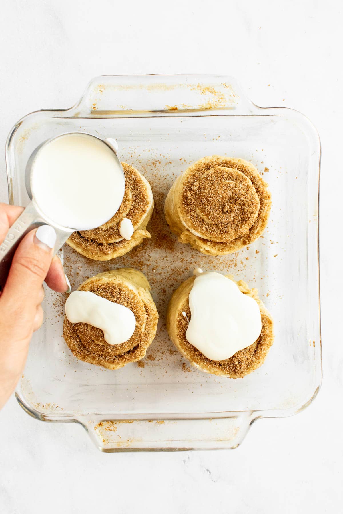 A hand pouring heavy cream from a measuring cup onto unbaked cinnamon rolls in a glass dish.