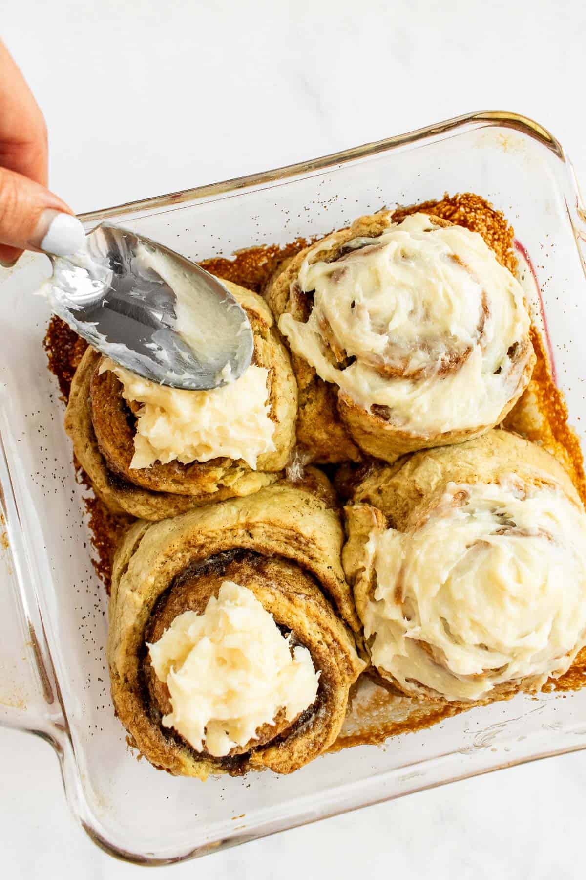 A hand spreading cream cheese frosting onto baked cinnamon rolls with the back of a spoon.