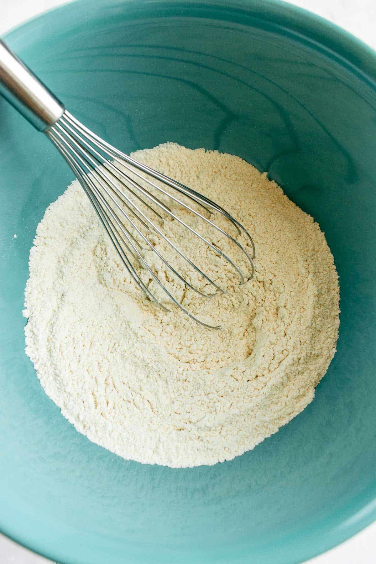 A blue mixing bowl of flour with a metal whisk resting in it.