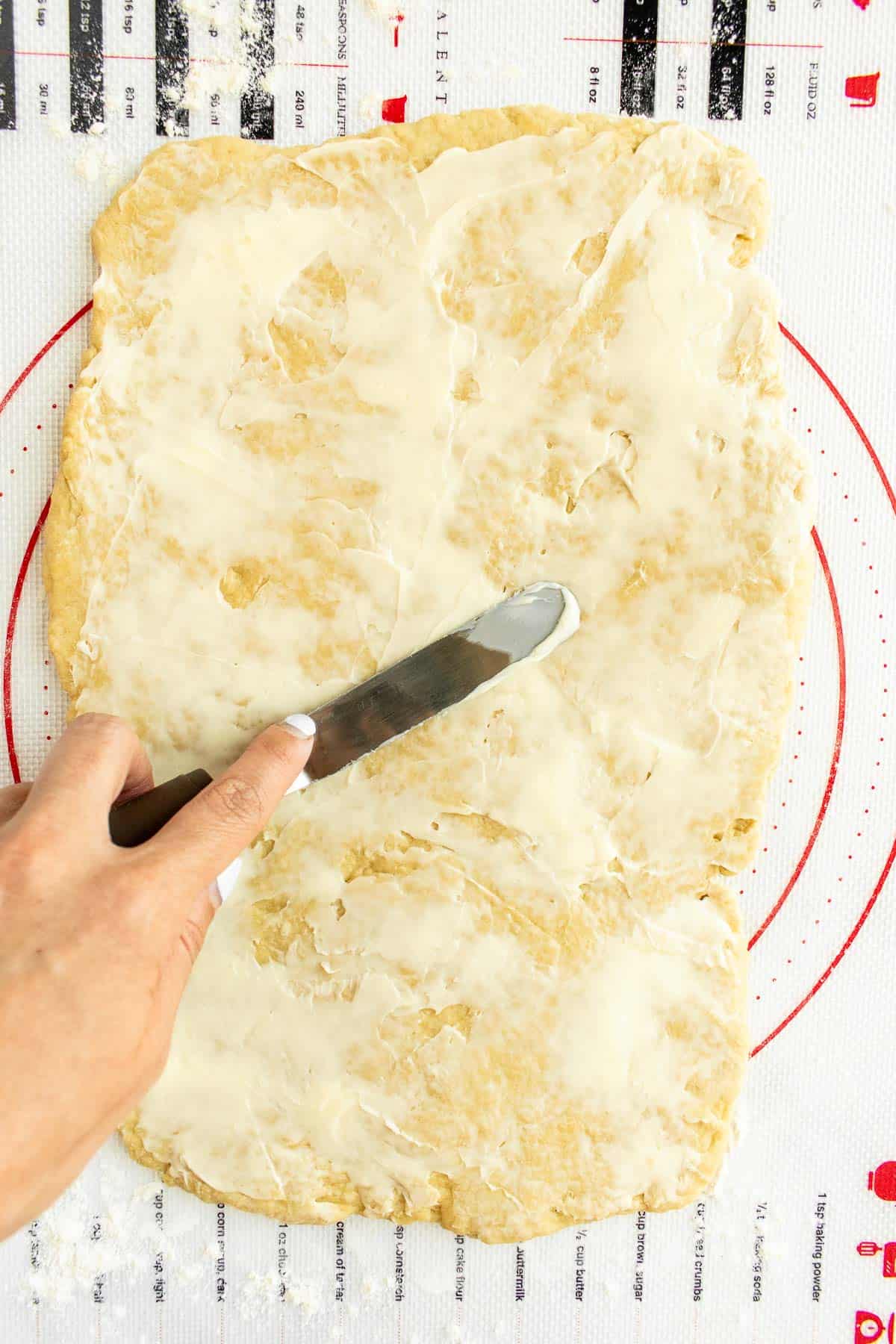 A hand spreading softened butter with a knife on rolled-out dough.