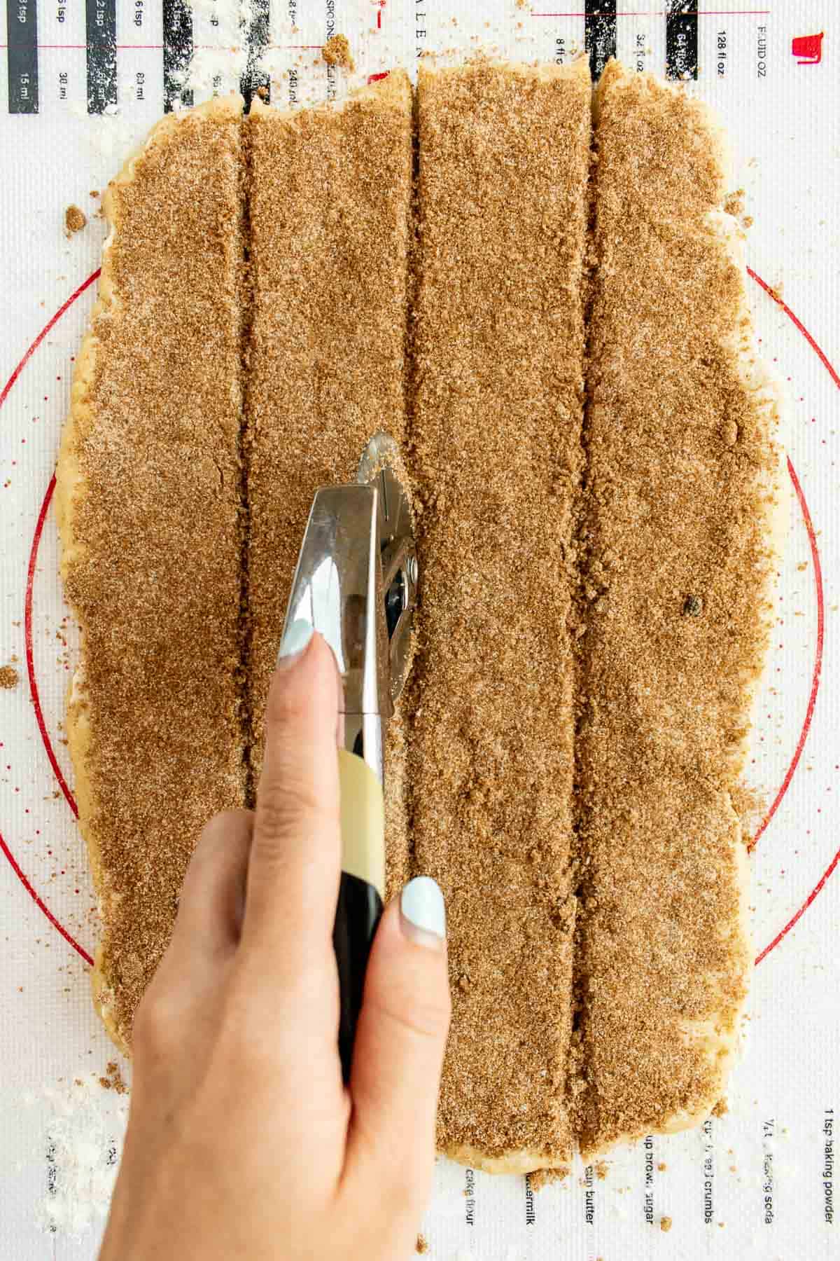 A hand using a pizza cutter to slice sugar-coated dough into four long strips.