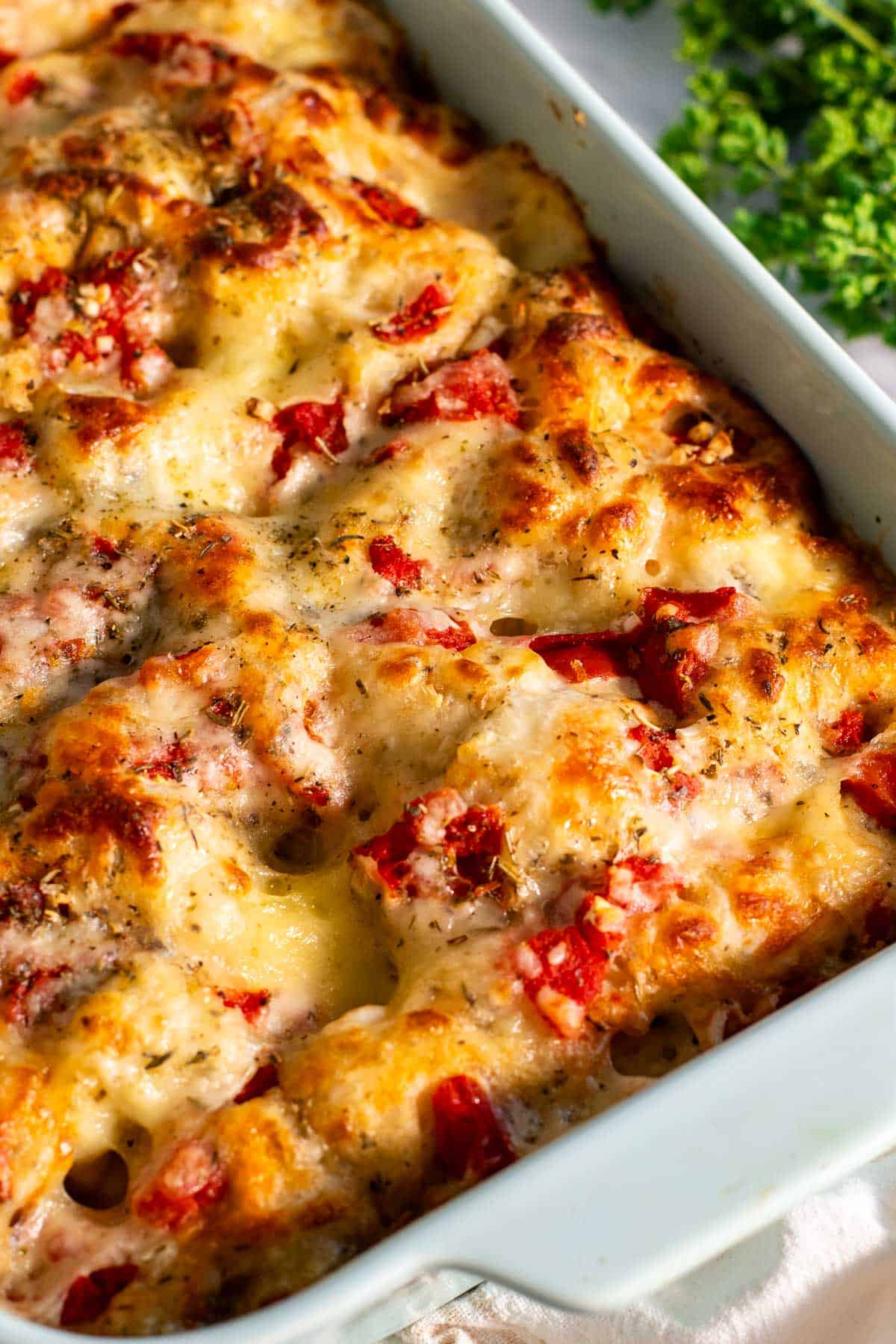 Baked focaccia pizza topped with melted cheese and diced tomatoes in a blue baking dish next to fresh oregano.