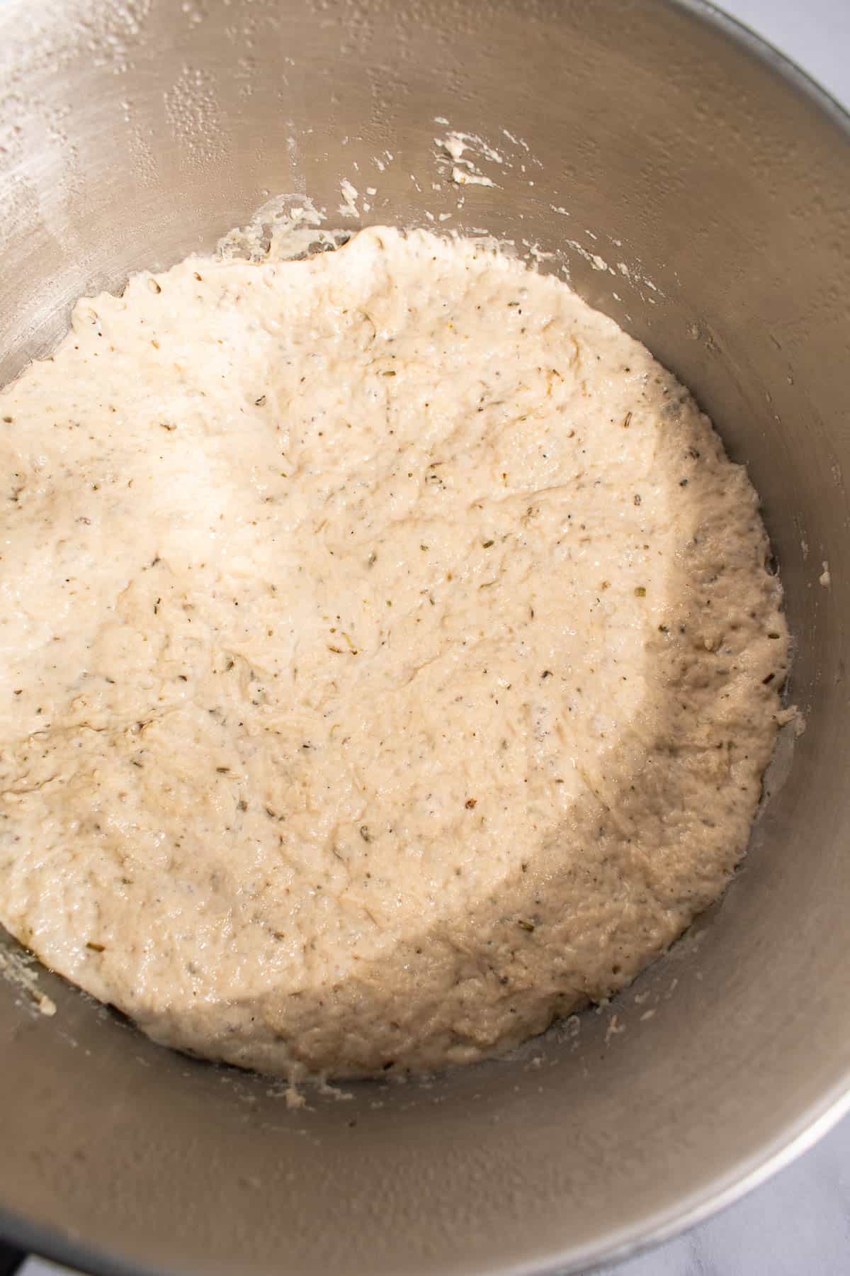 Risen, bubbly focaccia dough with dried oregano mixed in, in a metal bowl.