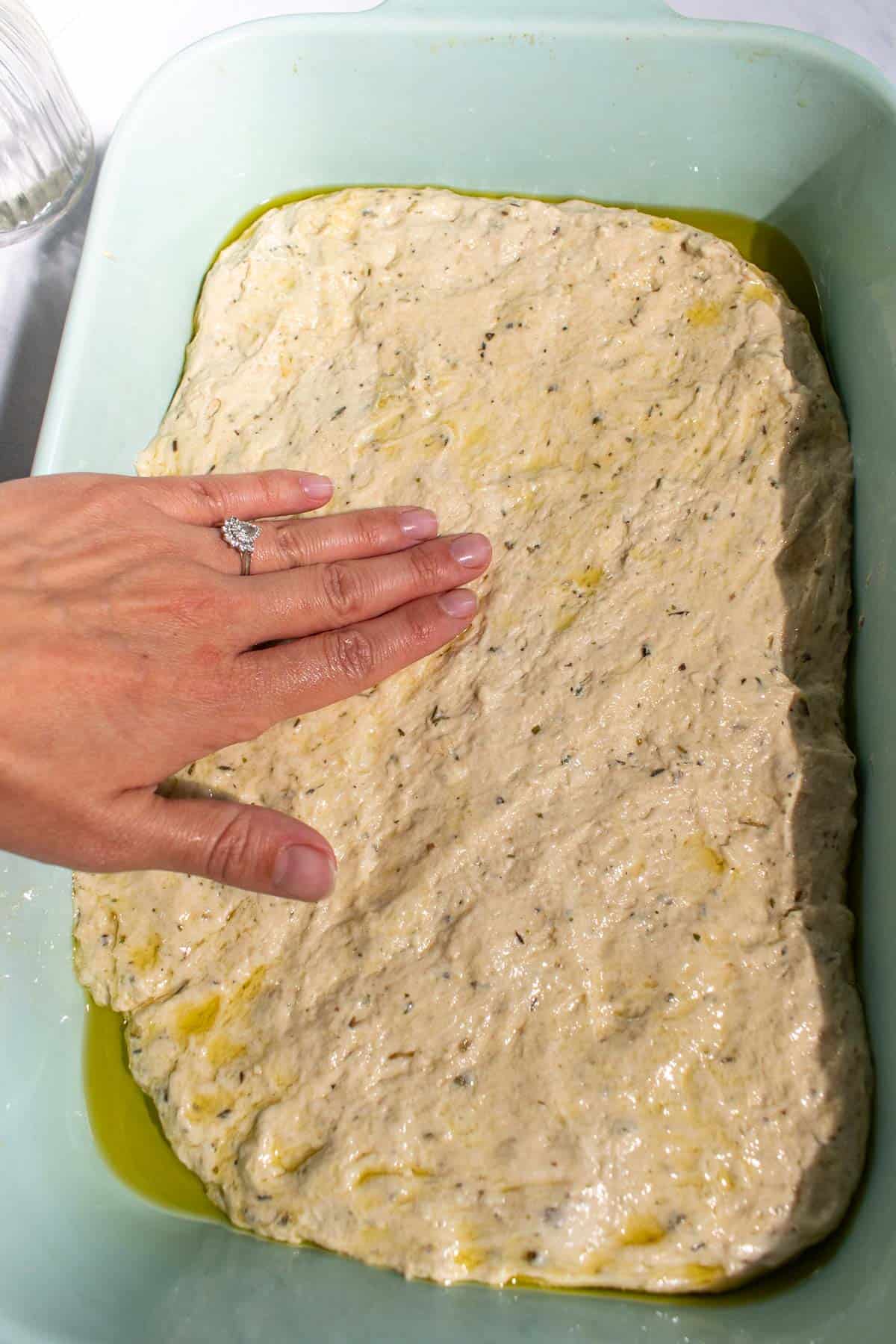 A hand spreading focaccia dough into a blue baking dish greased with olive oil.