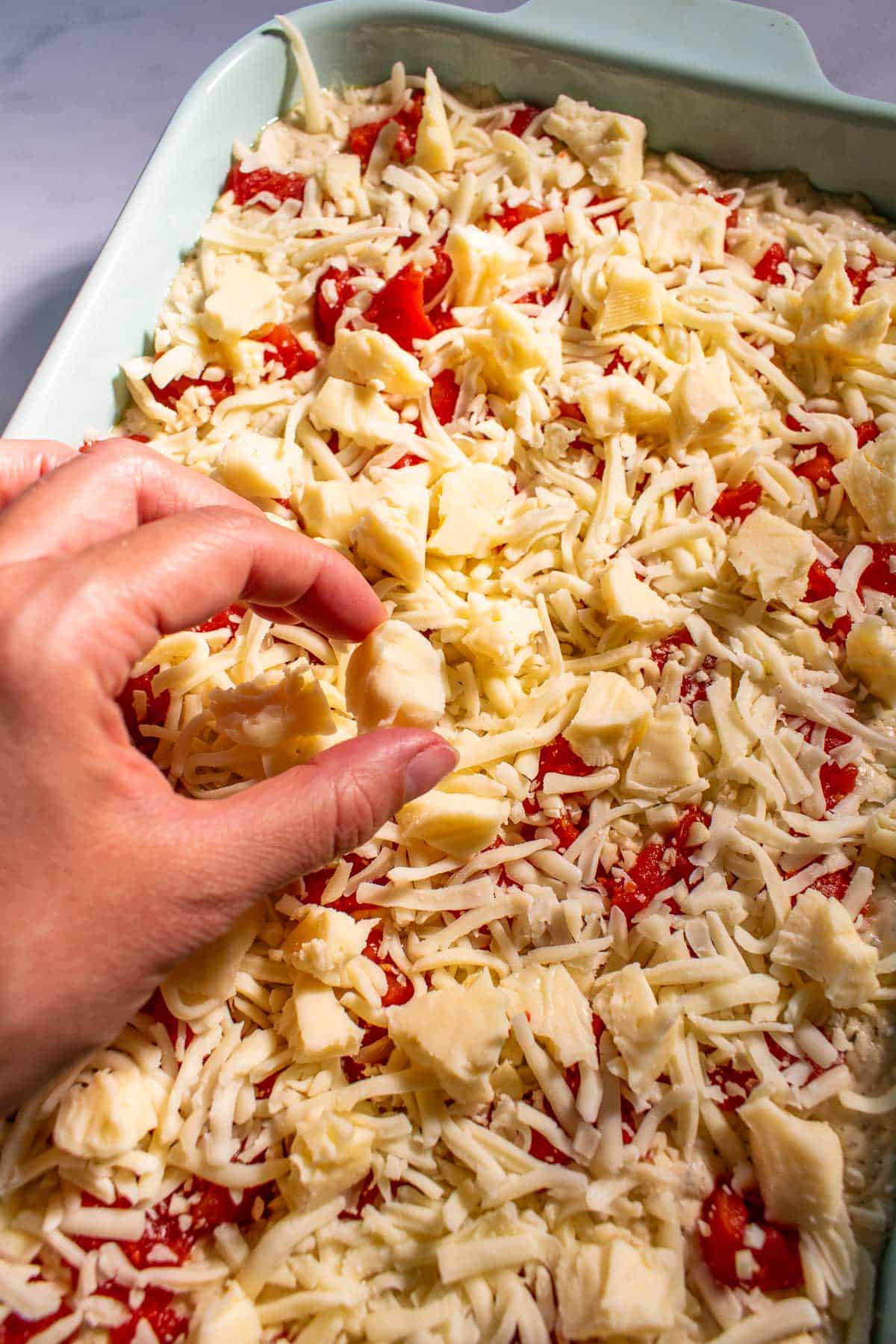 A hand placing a torn piece of cheese onto focaccia dough in a blue baking dish.