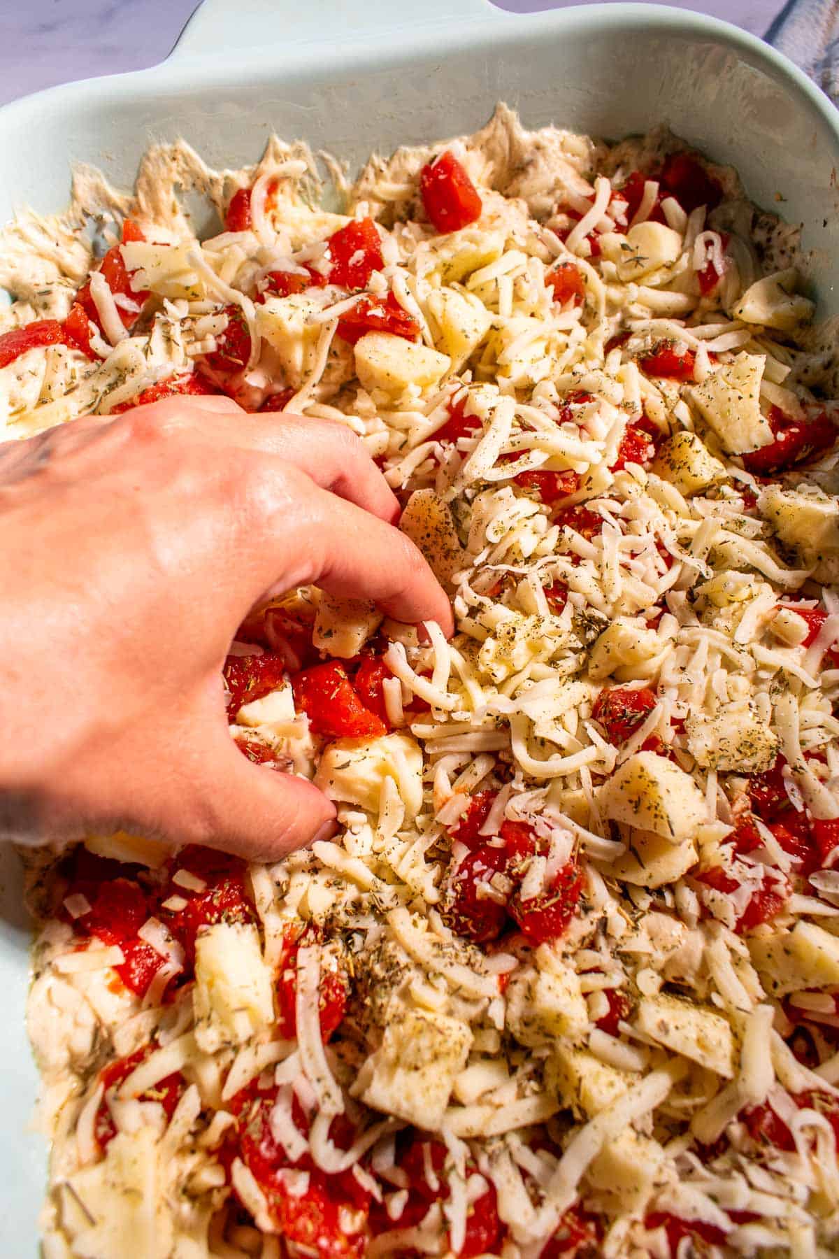 A hand dimpling focaccia dough topped with tomatoes and cheese in a large baking dish.