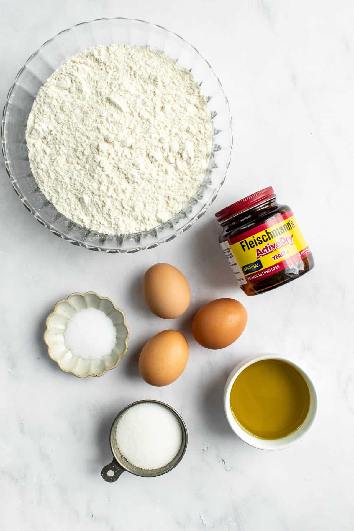 Three eggs in the middle of bowls of salt, sugar, olive oil, flour, and yeast on a light surface.