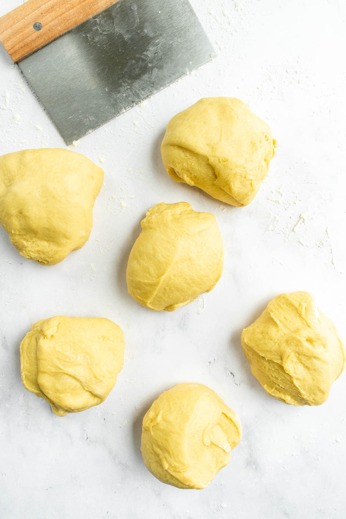Six pieces of bread dough on a light surface next to a metal bench scraper.