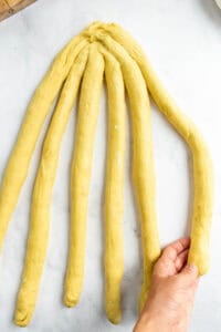 A hand picking up one strand of six bread dough strands laid out on a light surface.