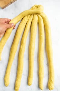 A hand crossing one strand over five other bread dough strands laid out on a light surface.