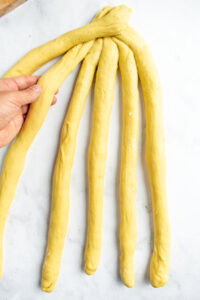 A hand braiding challah bread on a light surface with six long strands of dough.