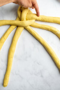 A hand pointing to a strand of bread dough that's being braided on a light surface.