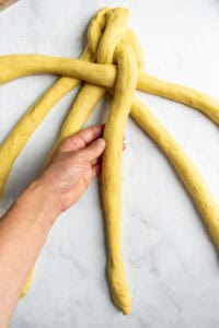 A hand braiding six strand of challah bread dough on a light surface.