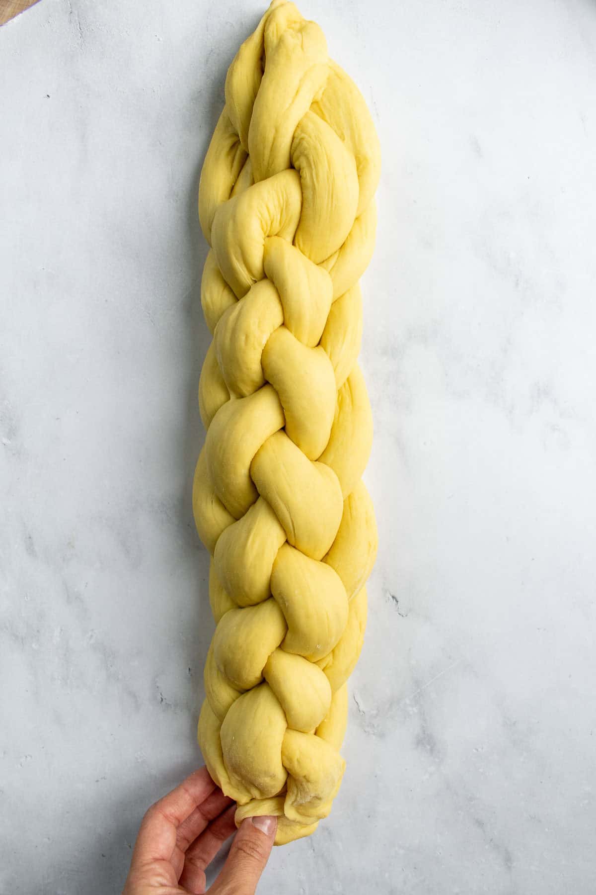 An unbaked six-stranded braided challah loaf on a light surface with a hand pinching the ends together.