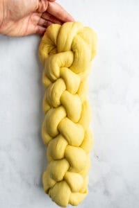 A hand tucking the edges of a braided loaf of six strand challah on a counter.