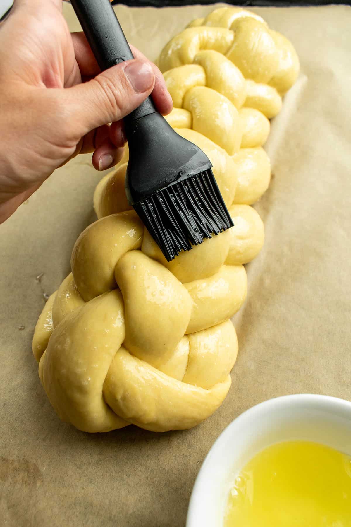 A hand brushing egg wash onto braided bread with a black silicone pastry brush.