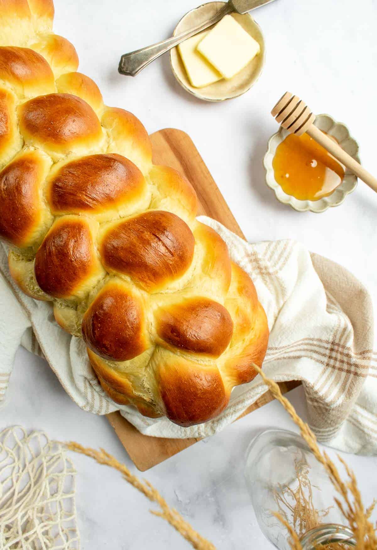A baked braided loaf of challah bread on a wood board next to small dishes of butter and honey.