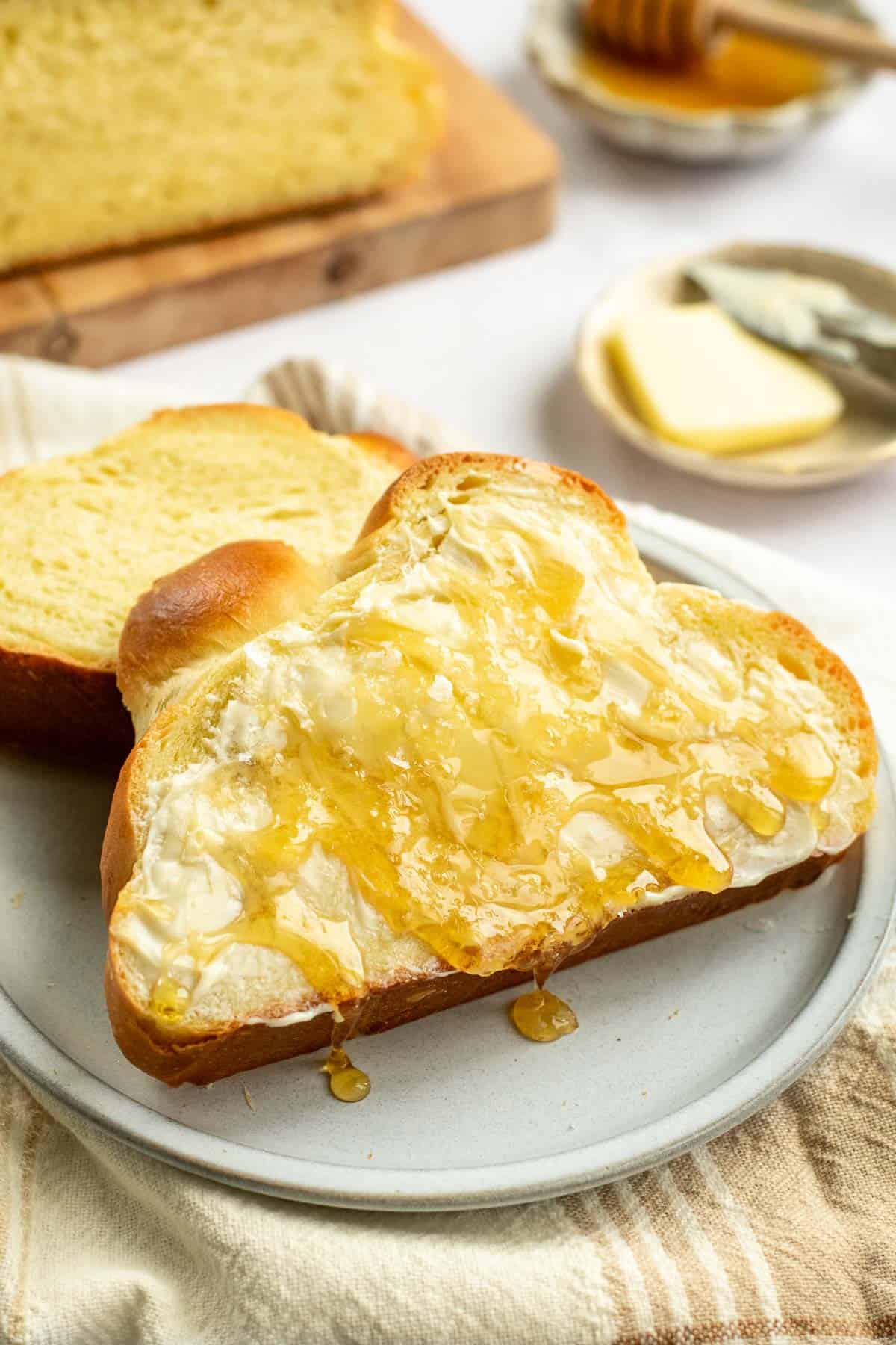 A slice of fluffy, soft challah bread on a grey plate with butter and honey spread on top.