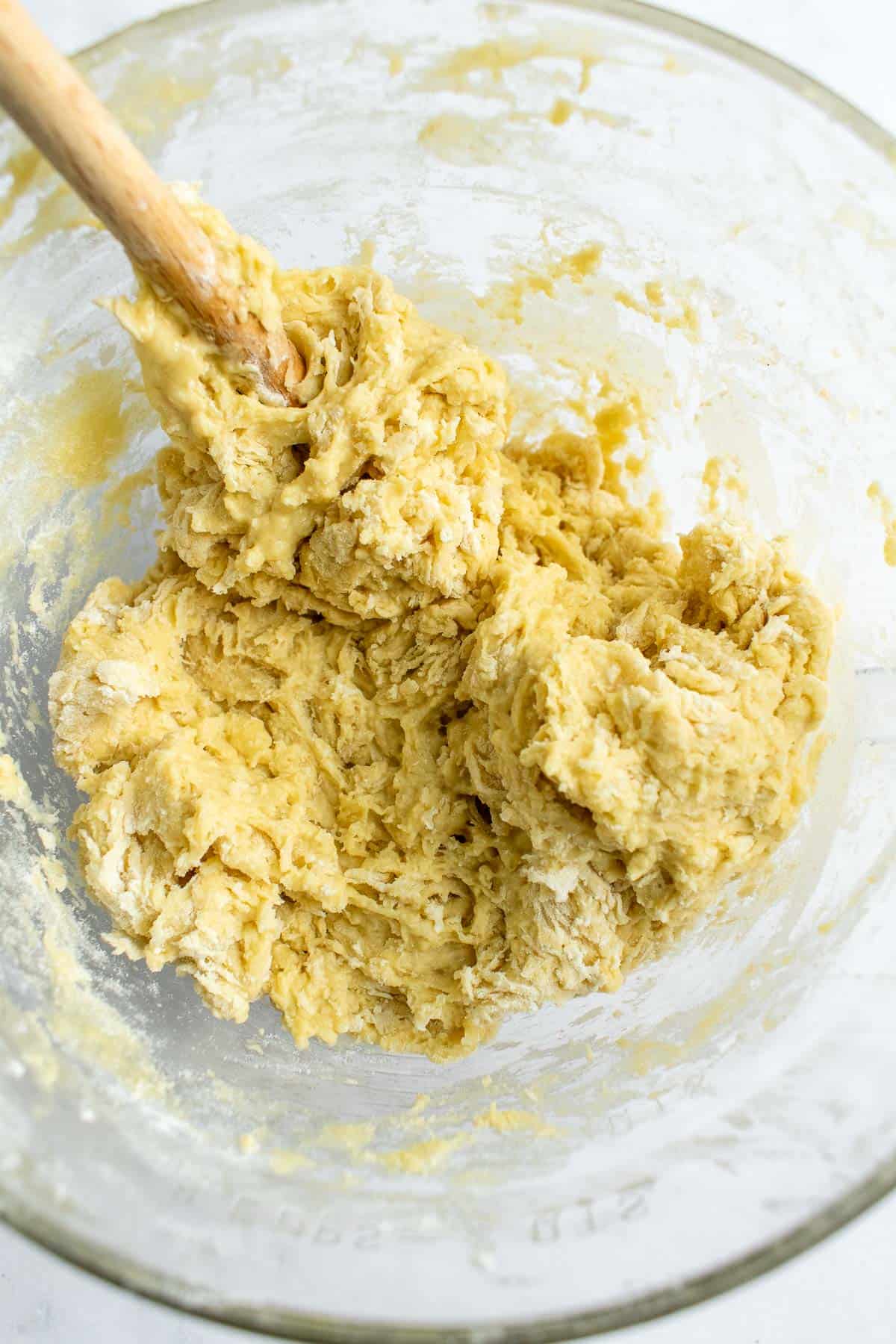 A shaggy, light yellow bread dough in a glass mixing bowl with a wooden spoon.
