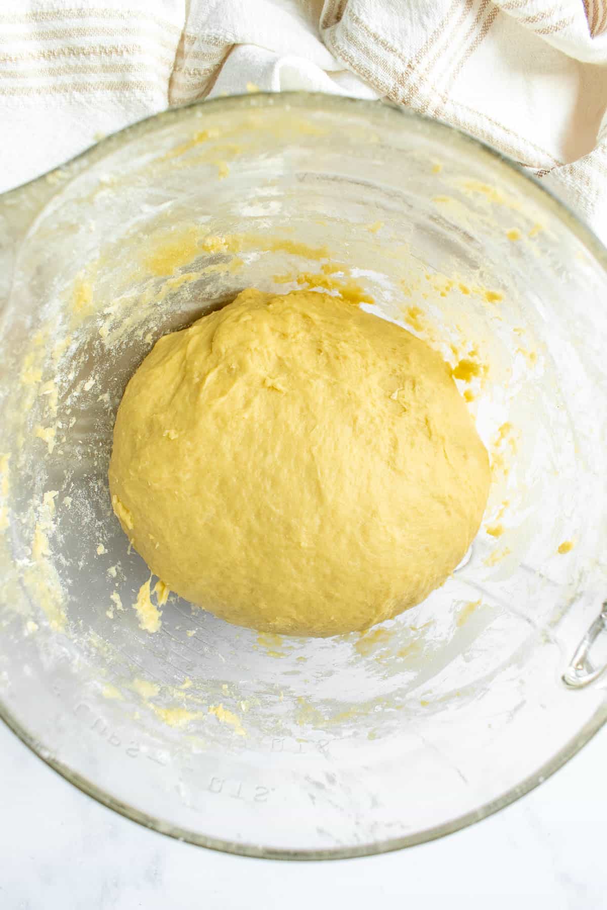 A ball of kneaded, light yellow bread dough in a glass mixing bowl next to a kitchen linen.