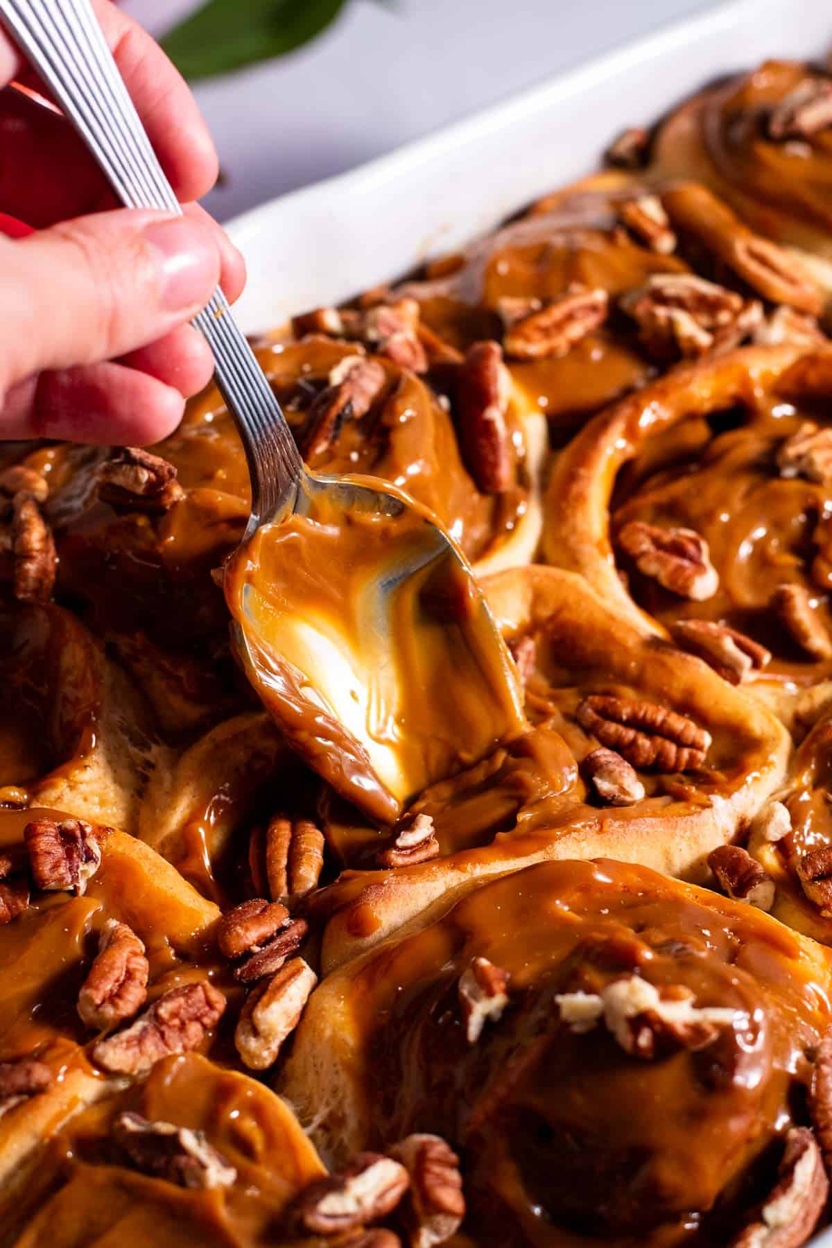 A hand spooning dulce de leche onto baked apple cinnamon rolls in a baking dish.