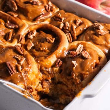 A white casserole dish filled with golden cinnamon rolls topped with dulce de leche with apples in the background.
