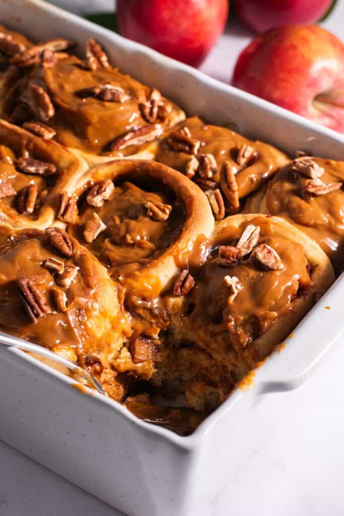 A white casserole dish filled with golden cinnamon rolls topped with dulce de leche with apples in the background.