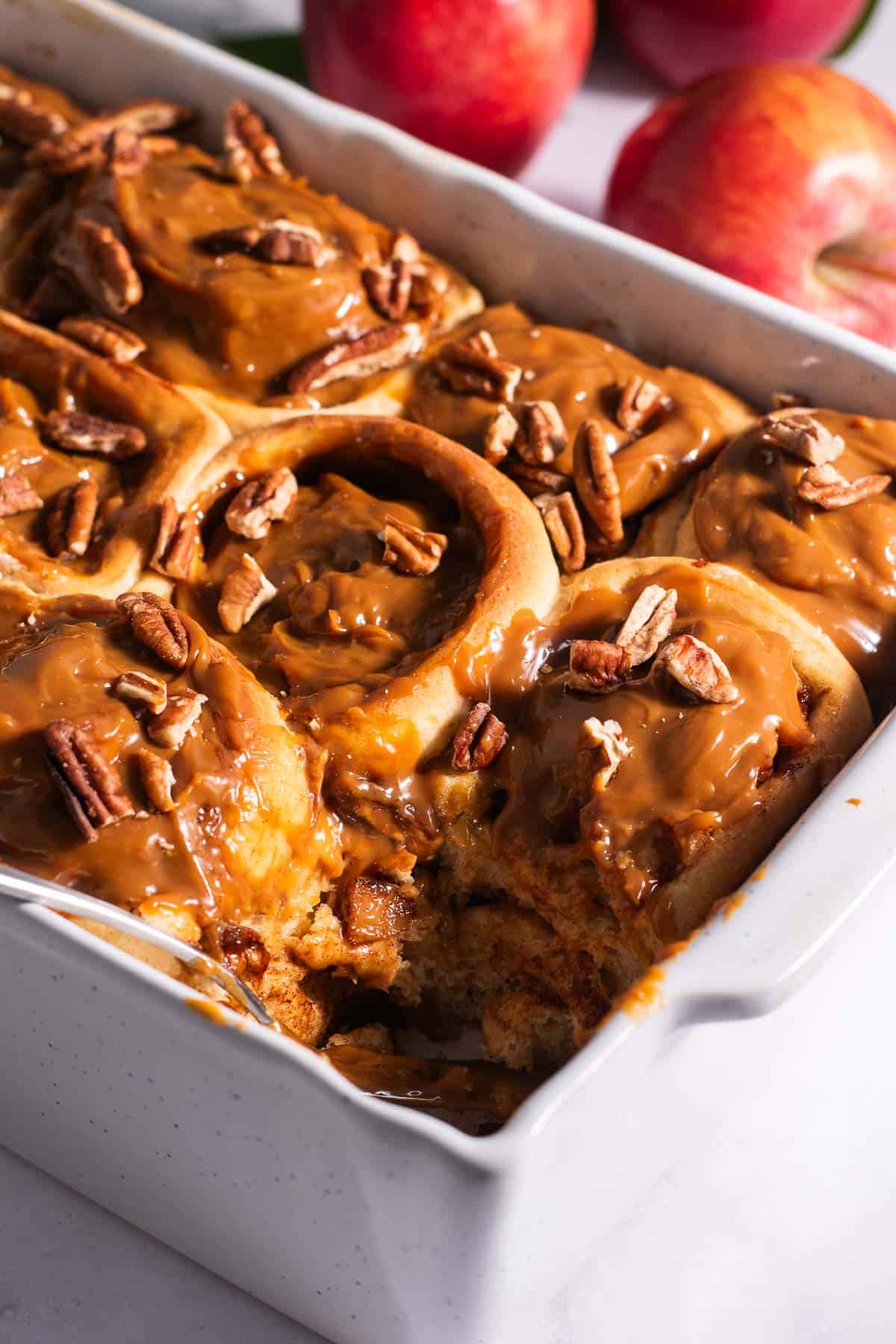 A white casserole dish filled with golden cinnamon rolls topped with dulce de leche with apples in the background.