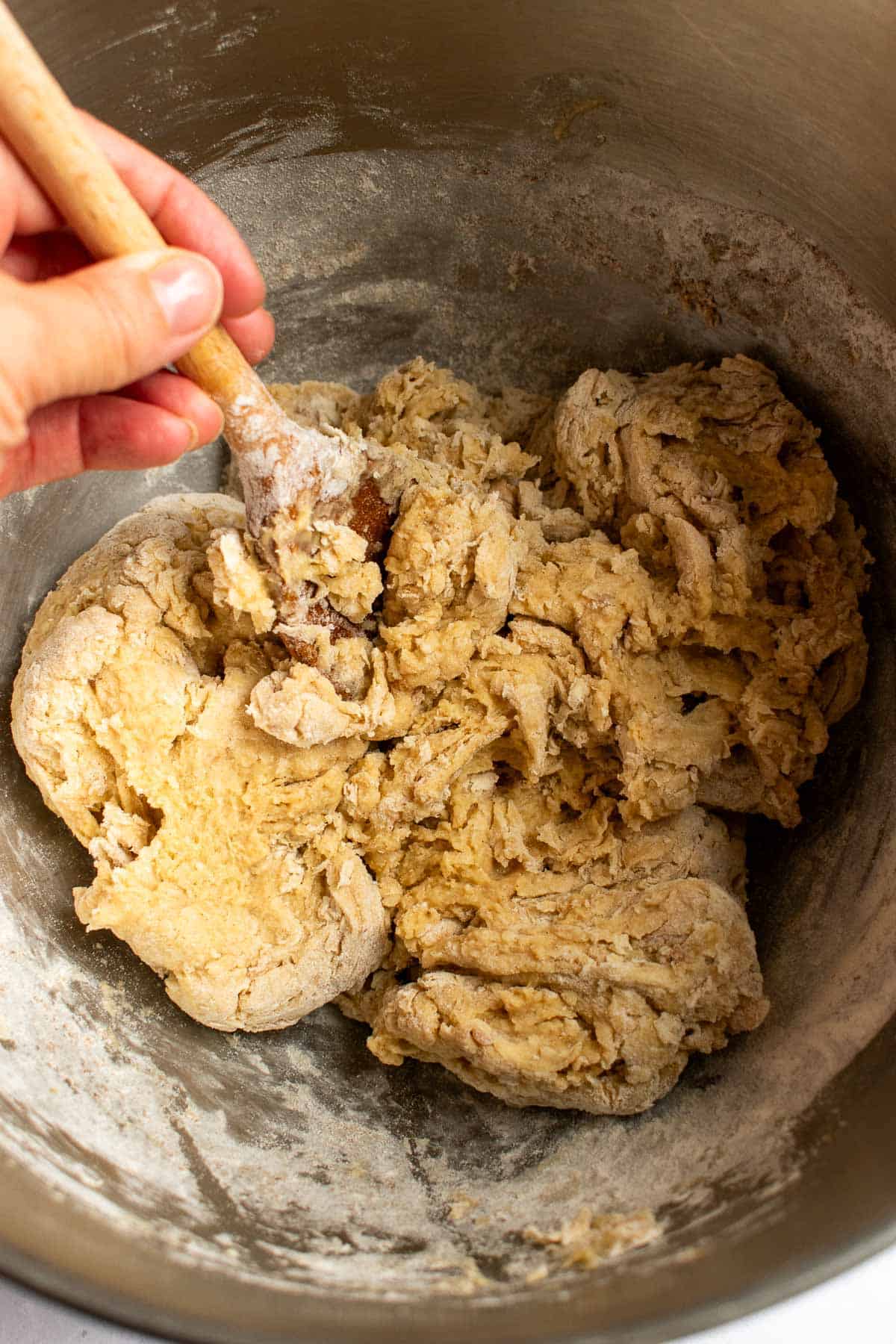 A hand mixing a shaggy bread dough in a metal bowl with a large wooden spoon.