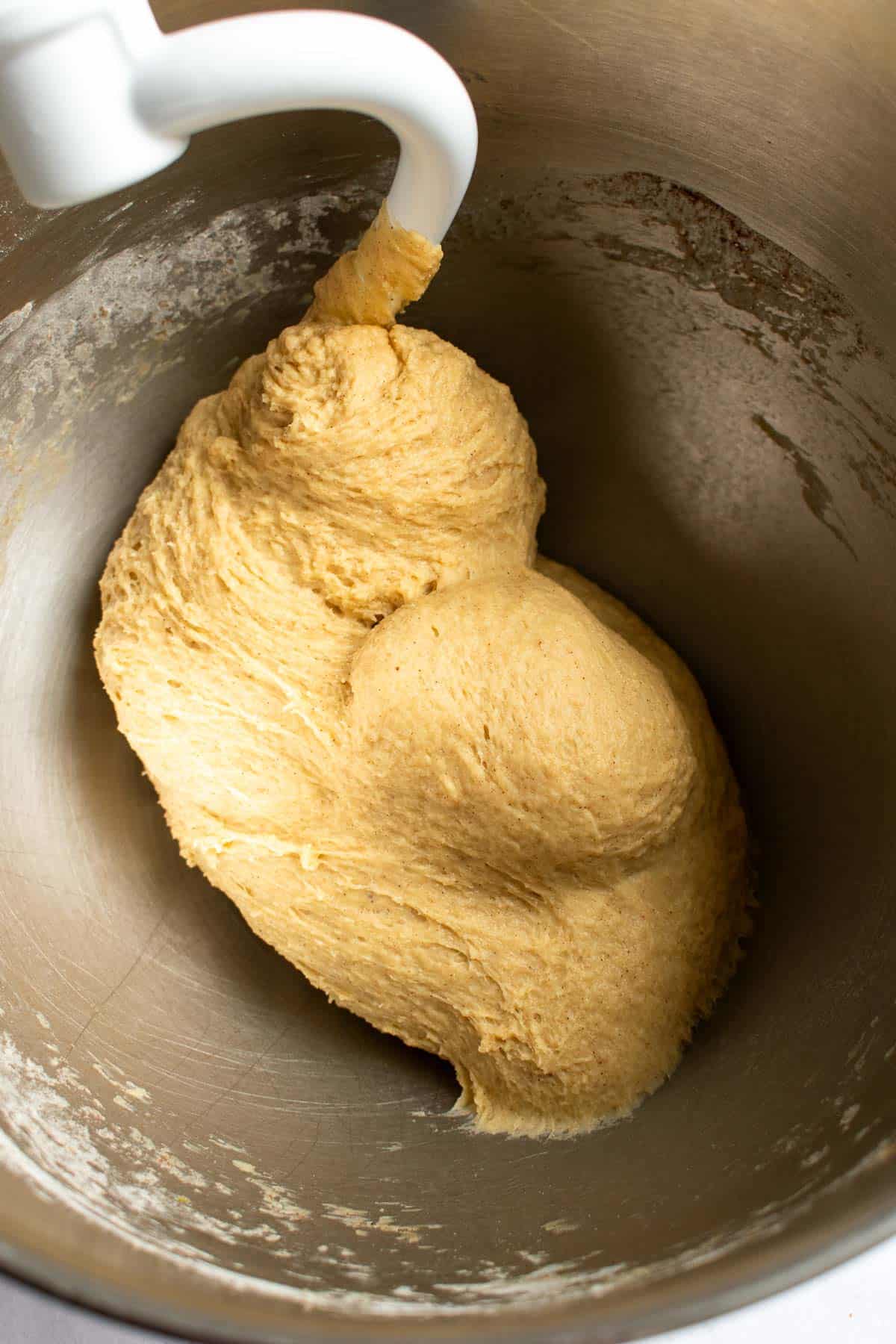 Shiny, kneaded cinnamon roll dough in a metal mixing bowl with a dough hook attachment from a stand mixer.
