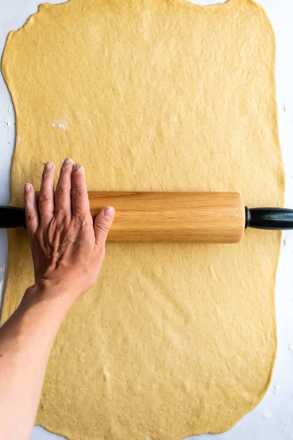 A hand rolling out cinnamon roll dough with a wooden rolling pin.
