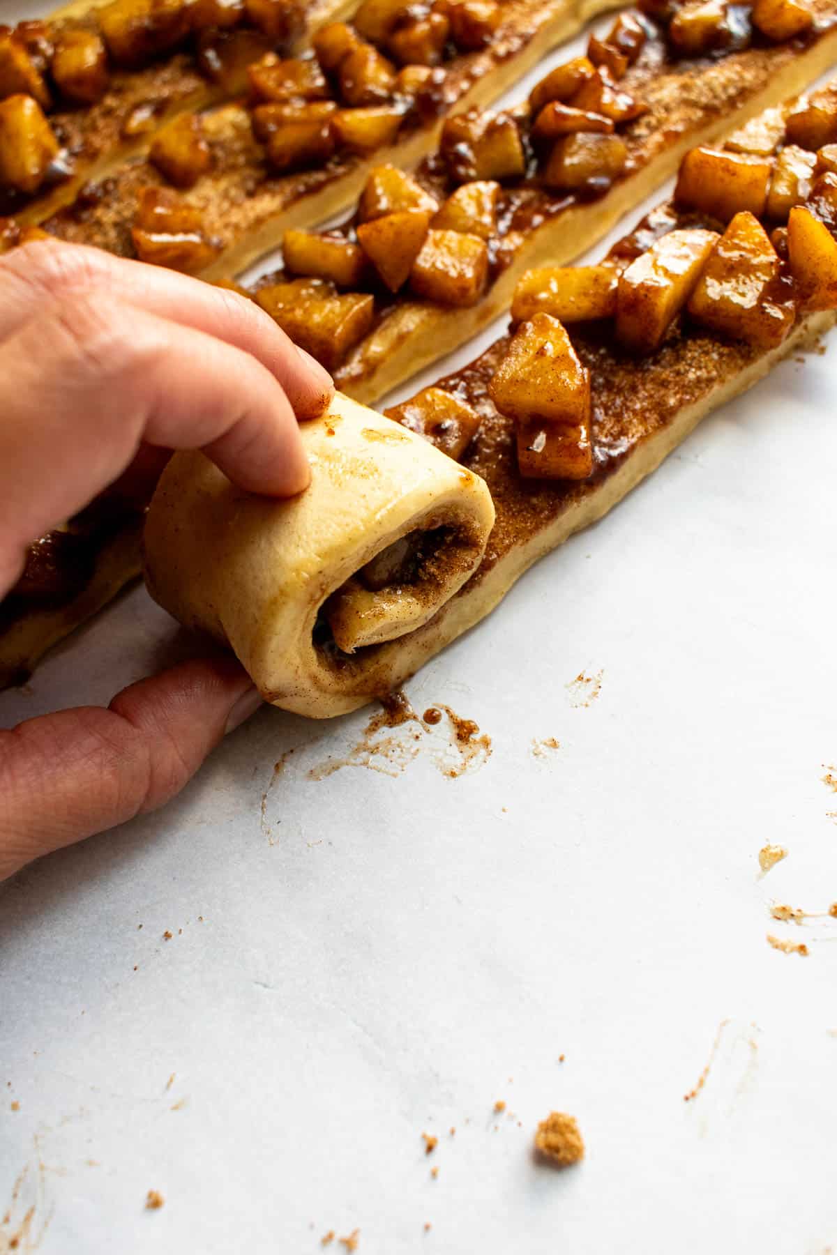 A hand rolling up a strip of dough around cooked apples and cinnamon sugar.