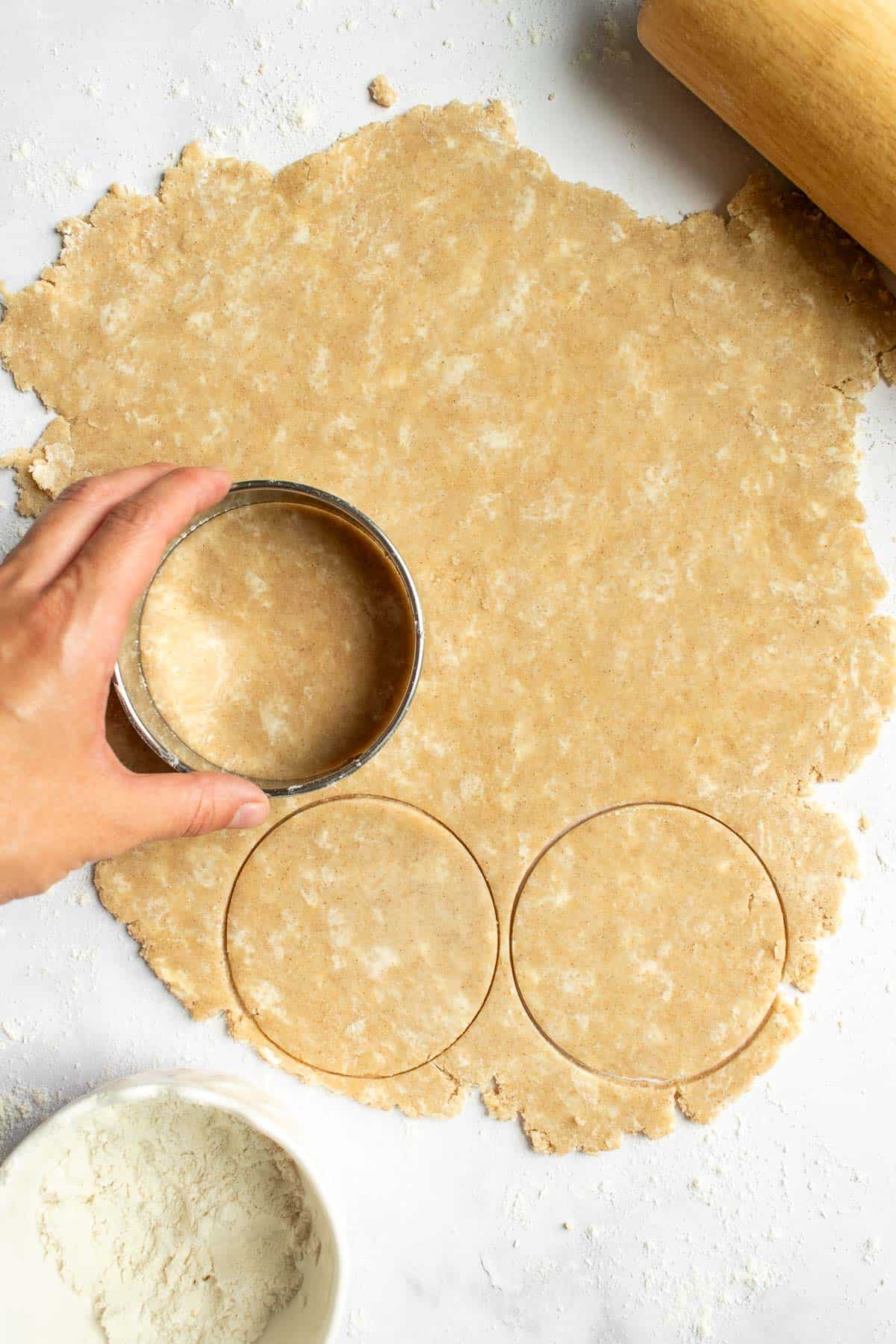 A hand cutting circles out of rolled-out pie crust next to a rolling pin.