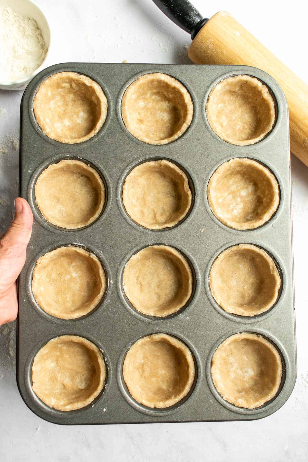 A hand holding a muffin tin filled with mini pie crusts.