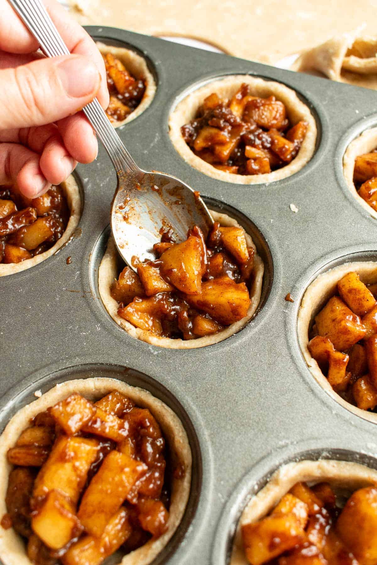 A hand spooning apple pie filling into mini pie crusts in a metal muffin tin.