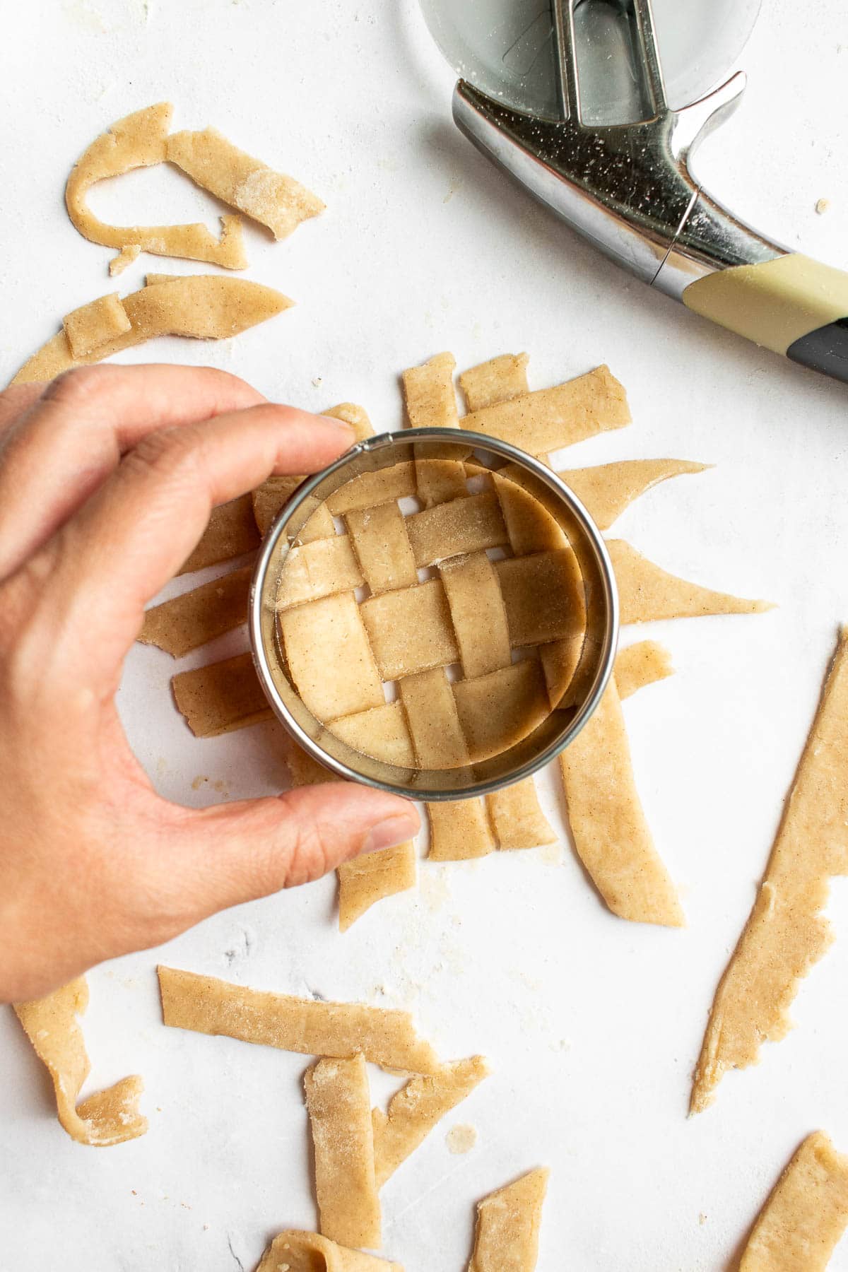 A hand cutting a circle with a cookie cutter from pie crust strips in a lattice design.