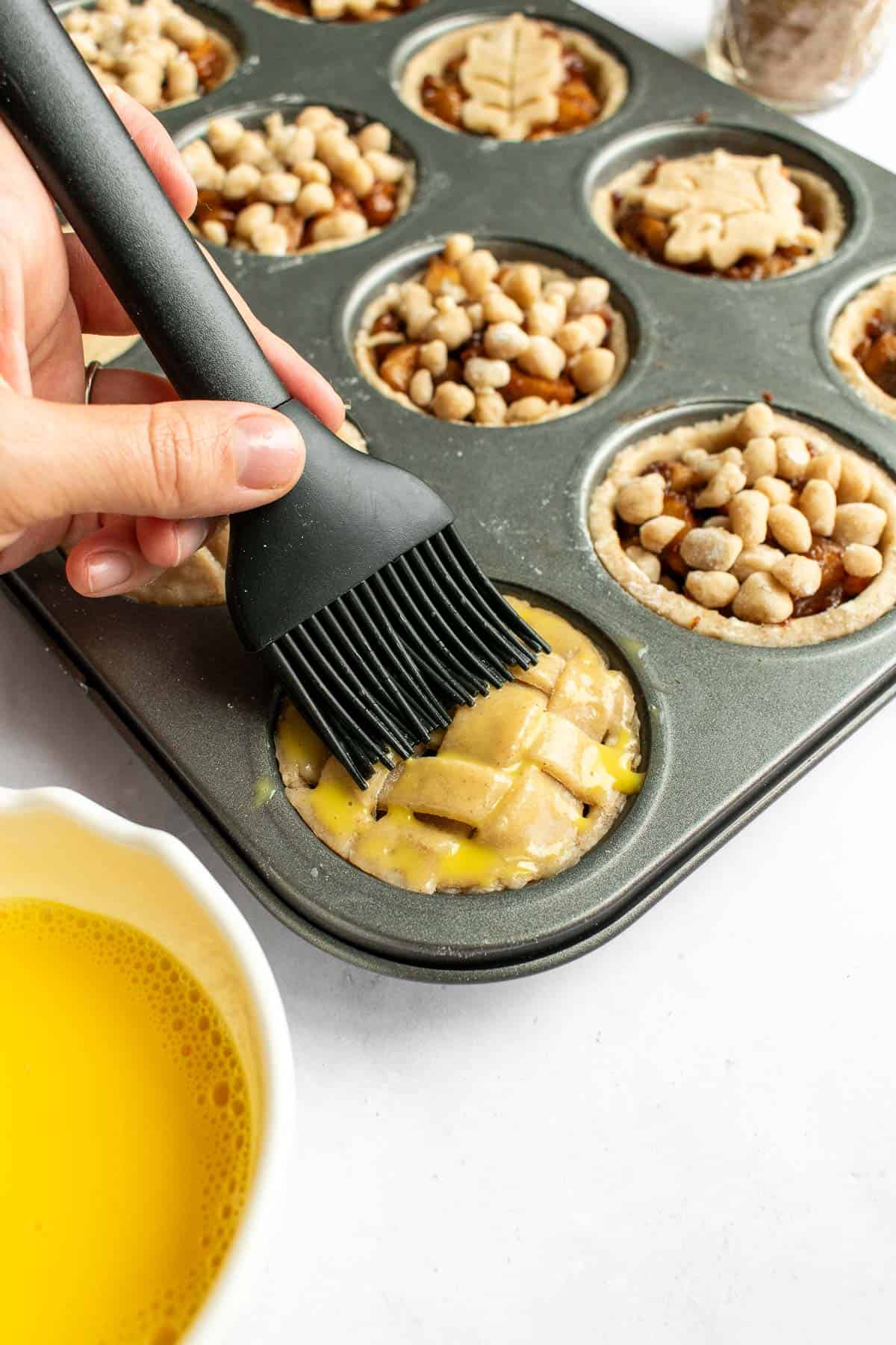 A hand brushing egg wash on top of mini apple pies in a metal muffin tin.