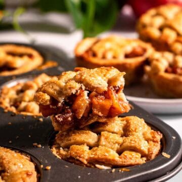 A muffin tin filled with mini pies, with a half-eaten mini apple pie sitting on top.