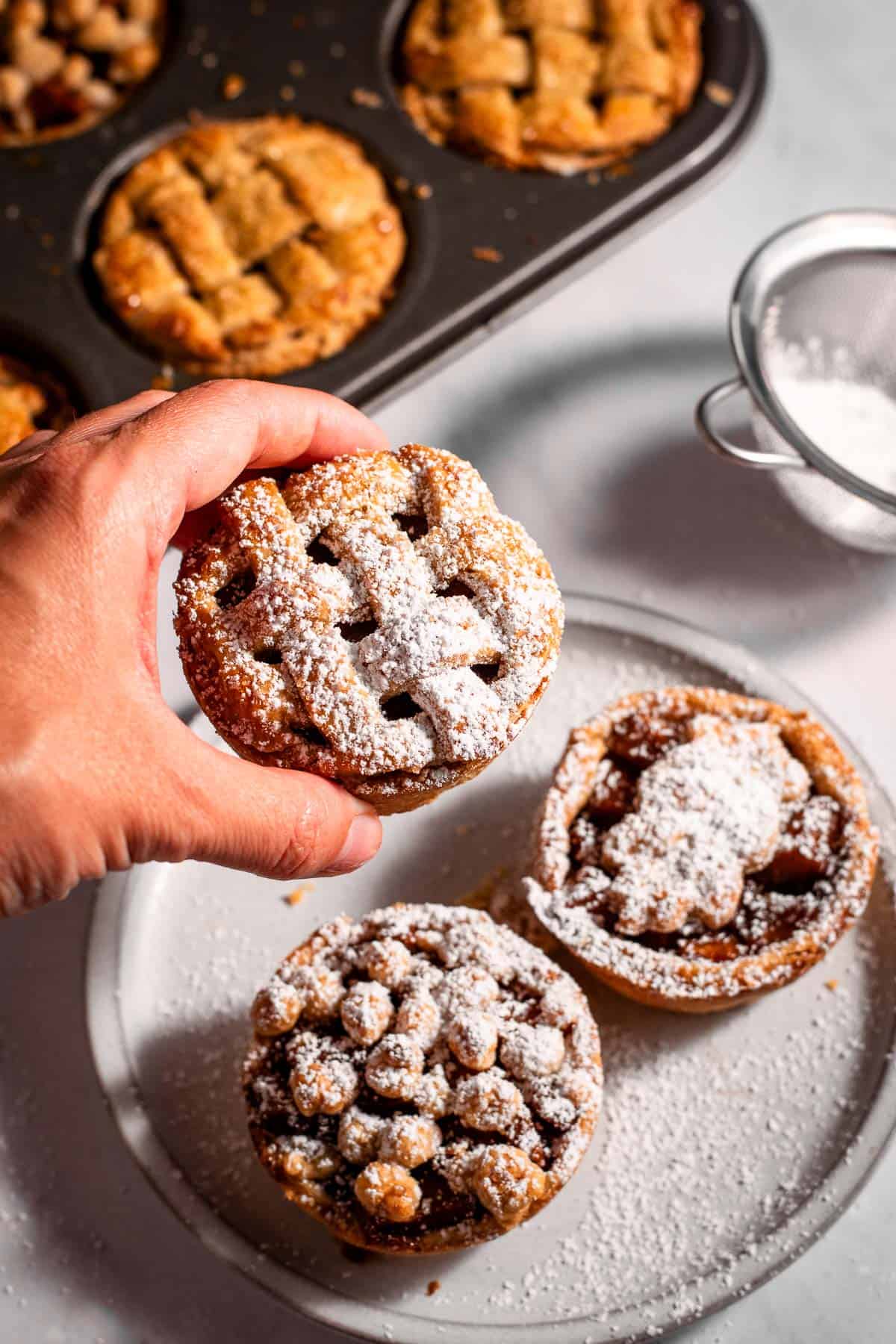 A hand holding a mini apple pie coated in powdered sugar next to more pies on a plate and in a muffin tin.