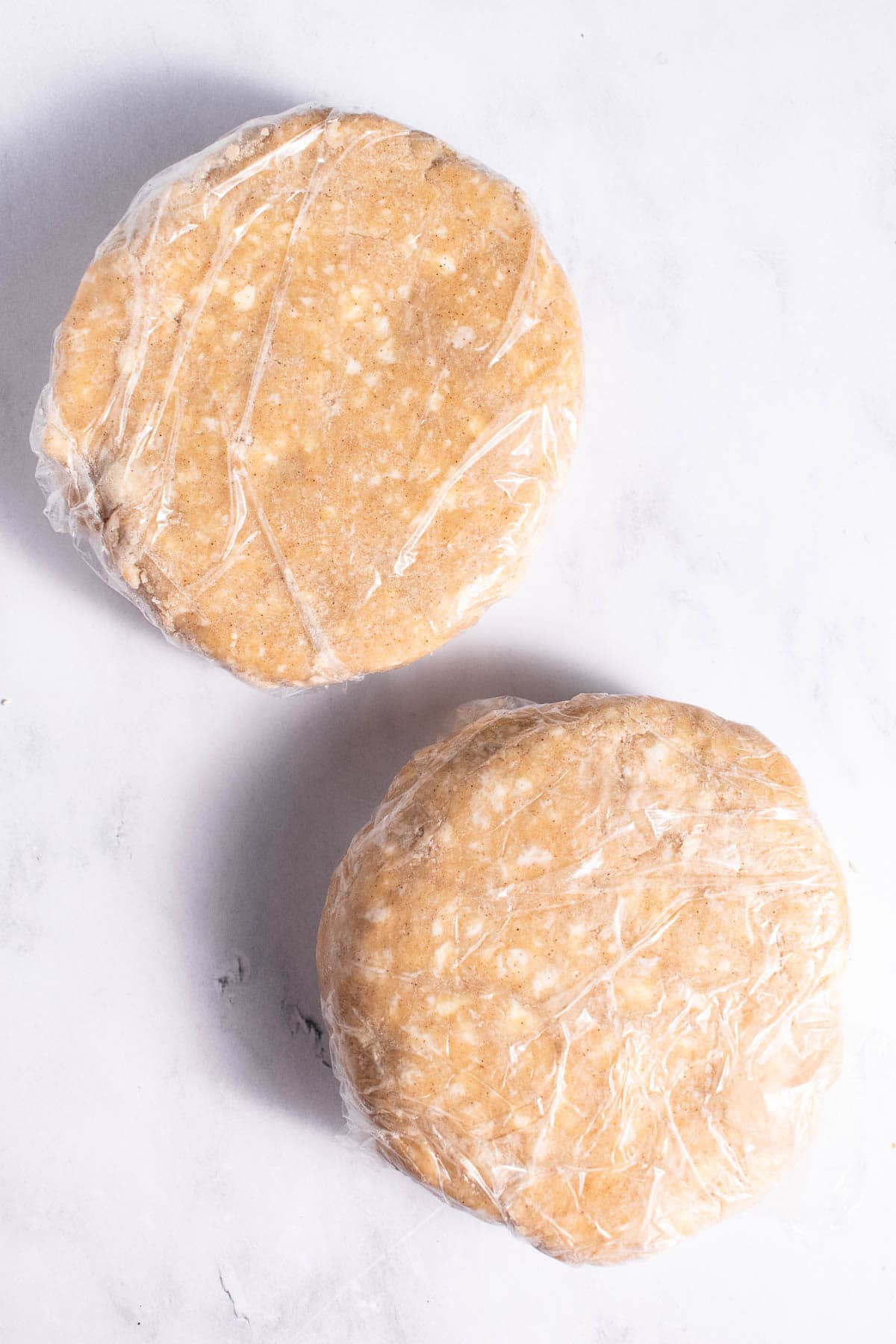 Two discs of pie crust wrapped in plastic wrap on a light counter.