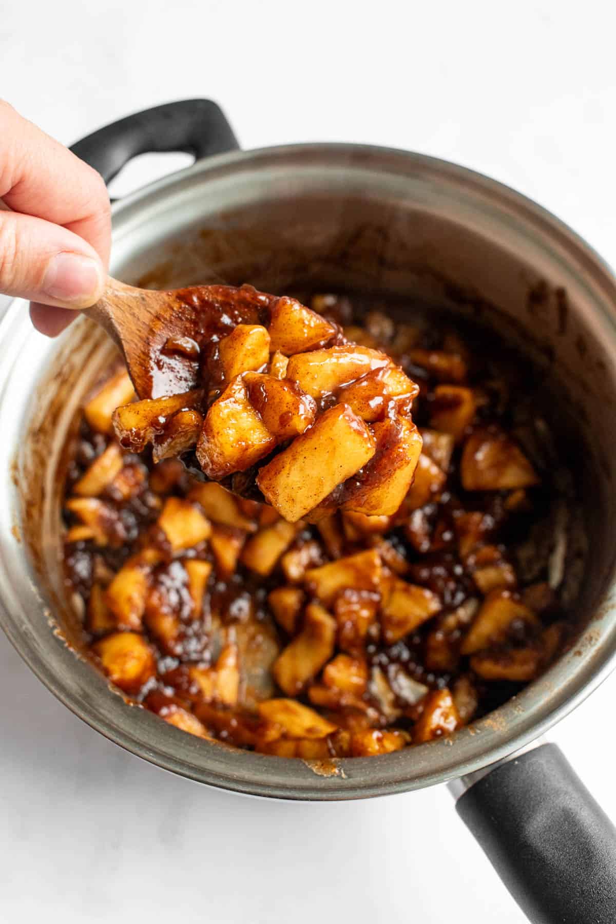 A hand holding a large spoon with caramelized apples over a metal saucepan.