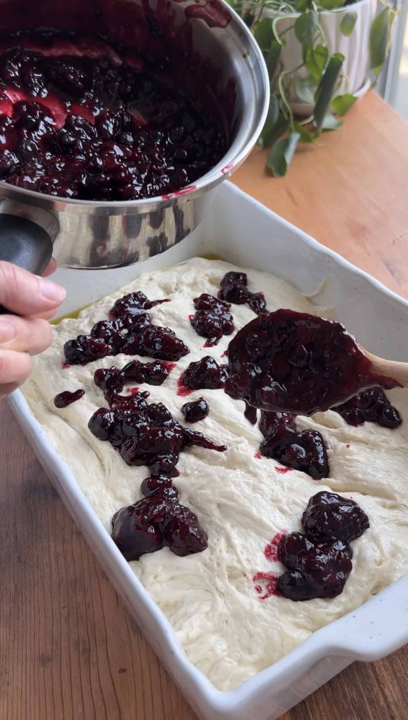 A hand spooning dollops of cooked berries onto focaccia dough in a baking dish.