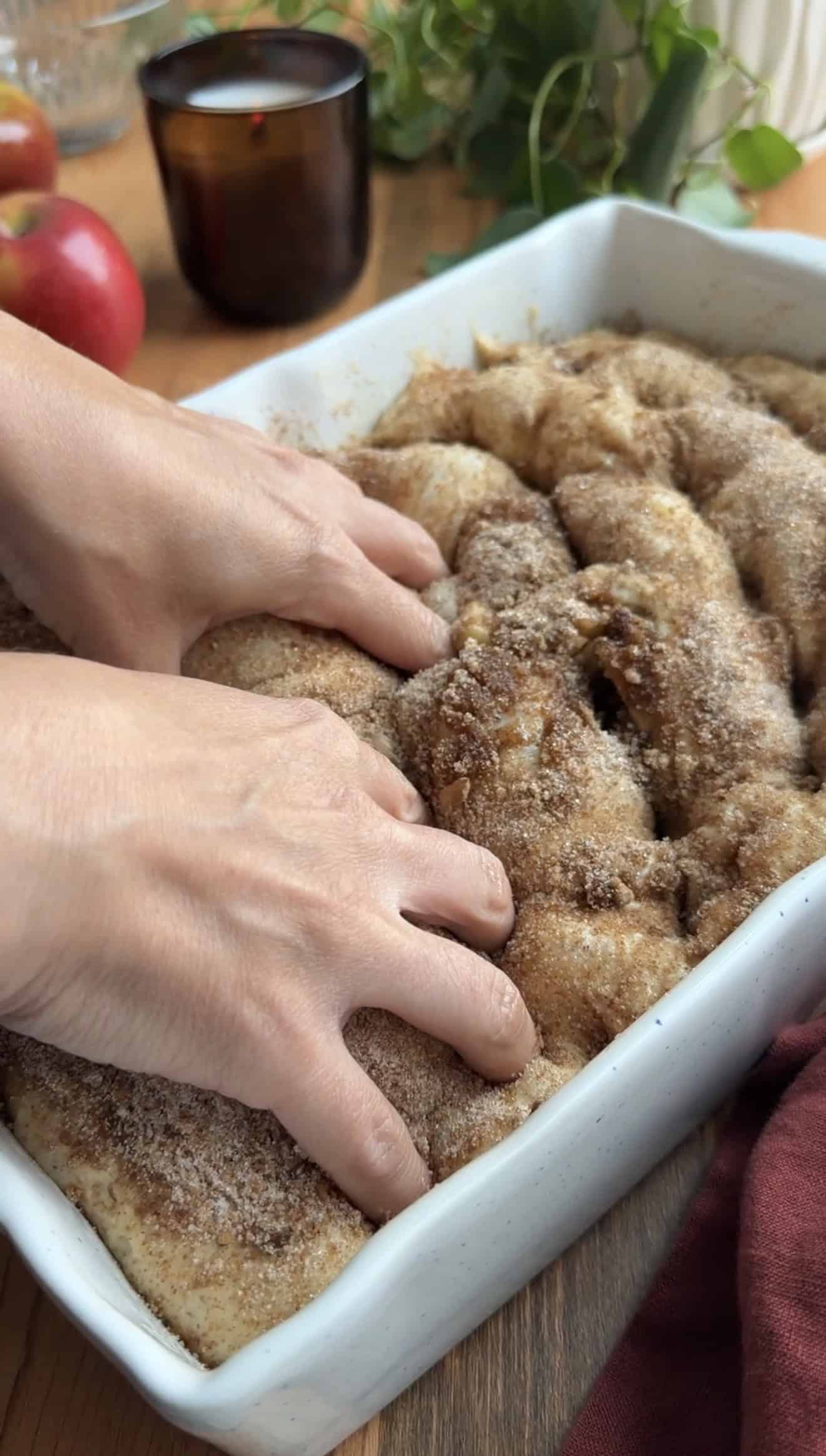 Two hands dimpling focaccia dough that's topped with cinnamon sugar in a baking dish.