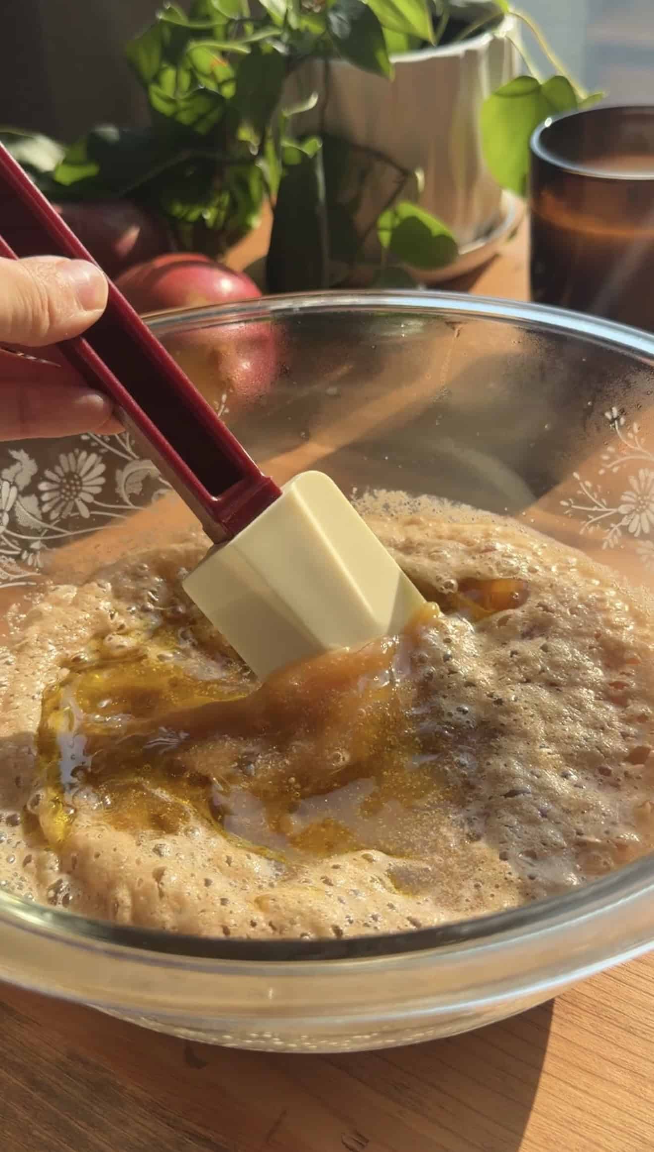 A hand stirring foamy yeast in apple juice in a large glass bowl with a spatula.
