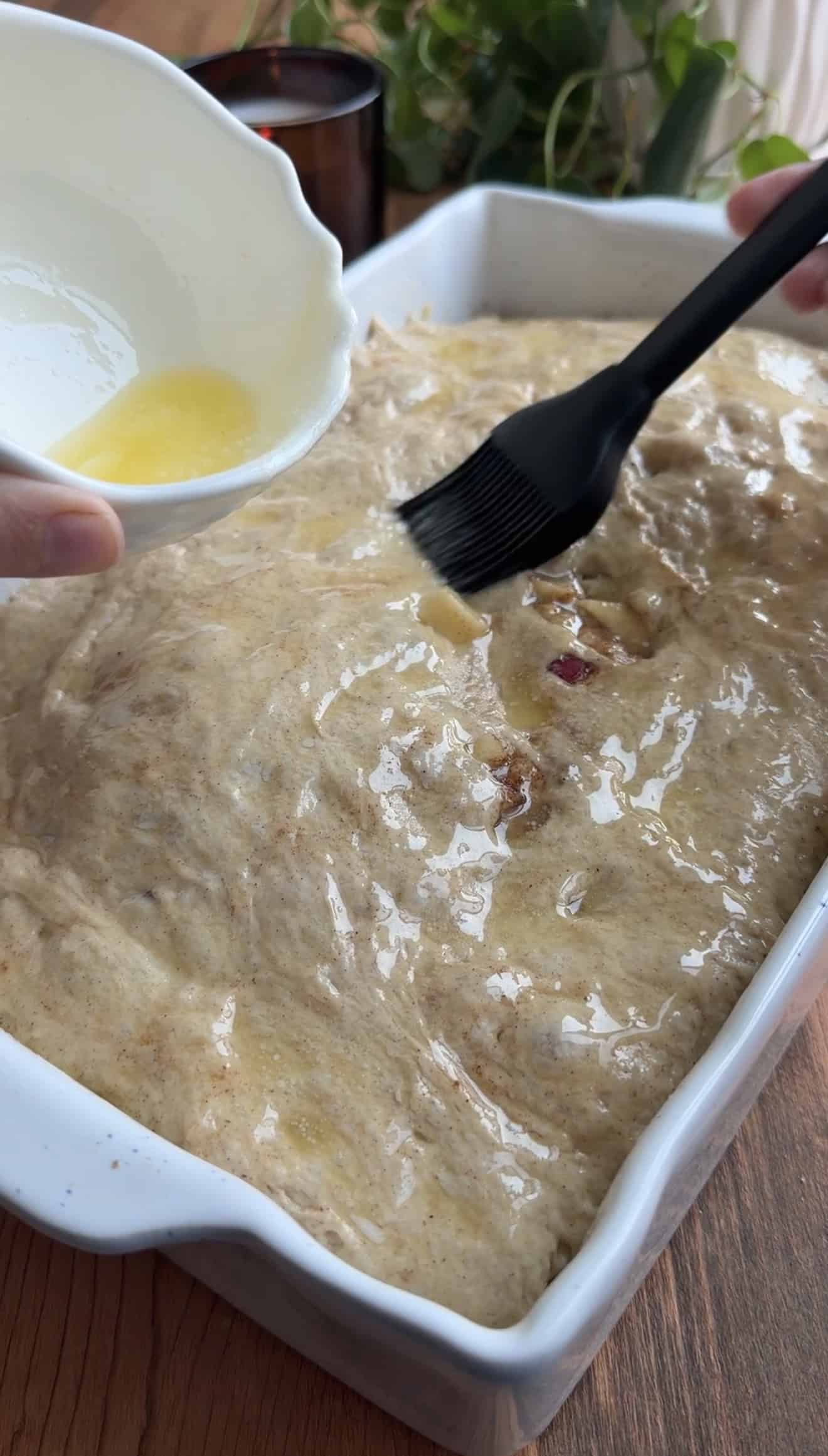 A hand brushing melted butter over the top of focaccia dough in a baking dish with a silicone pastry brush.